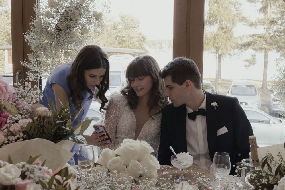 Wedding guests huddle together looking at a phone during a reception dinner with floral arrangements on the table.