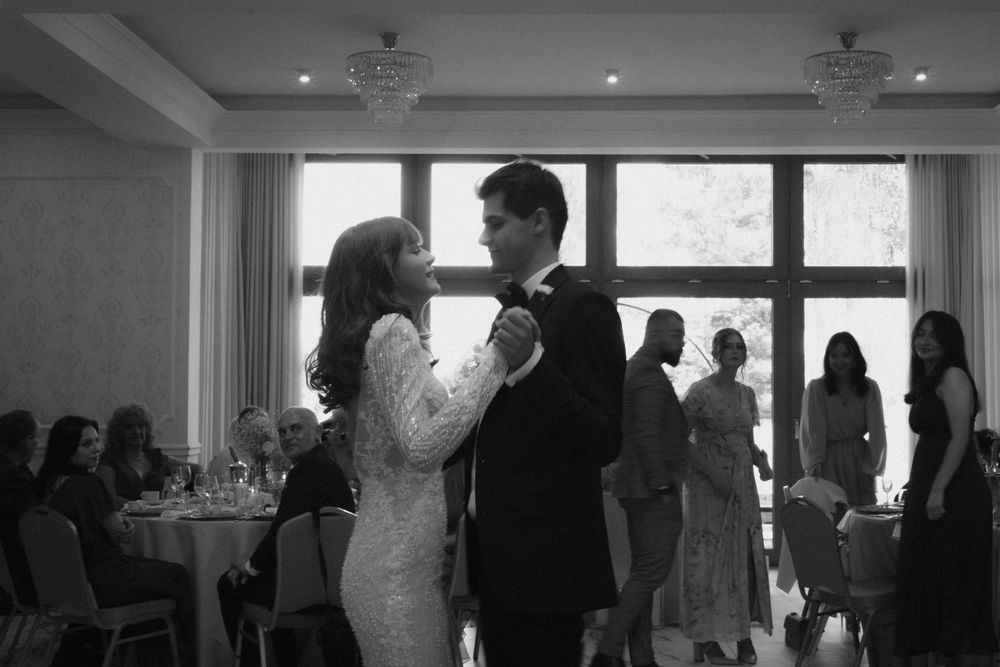 A couple shares their first dance during a wedding reception in a bright ballroom with guests seated at round tables.