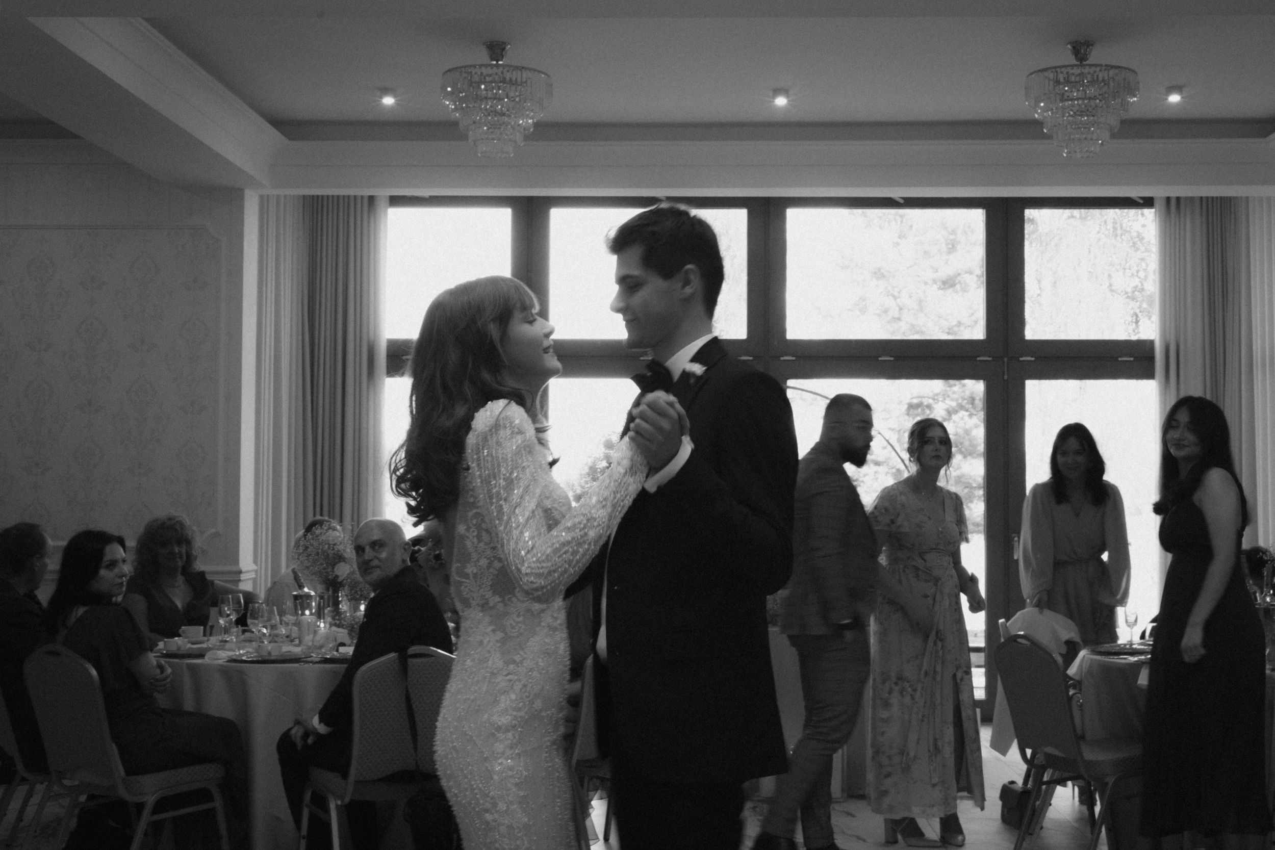 A couple shares their first dance during a wedding reception in a bright ballroom with guests seated at round tables.