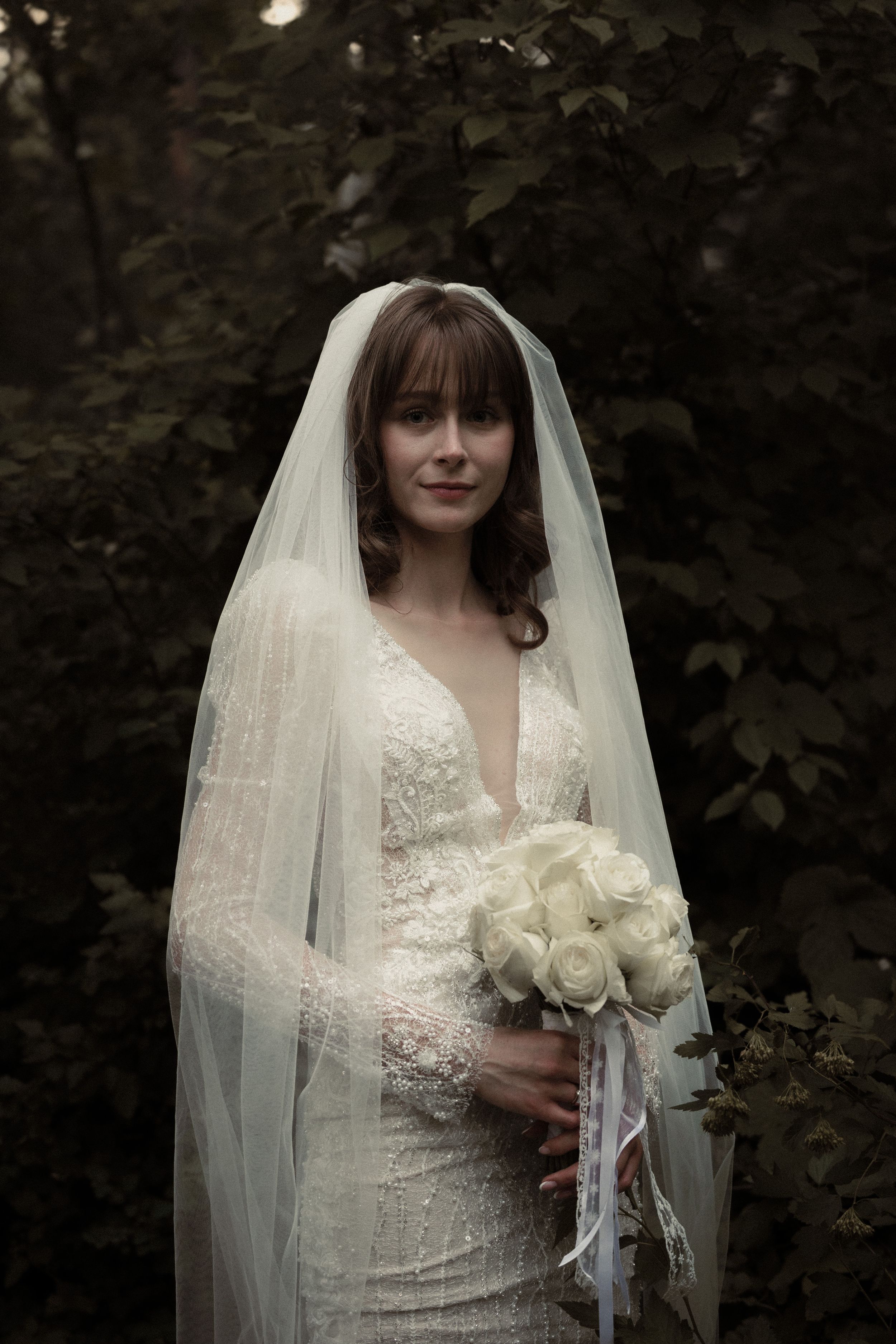 A bride in a fitted lace wedding dress with plunging neckline holds a white rose bouquet in a moody forest setting.