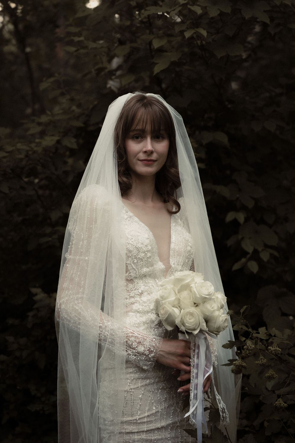 A bride in a fitted lace wedding dress with plunging neckline holds a white rose bouquet in a moody forest setting.