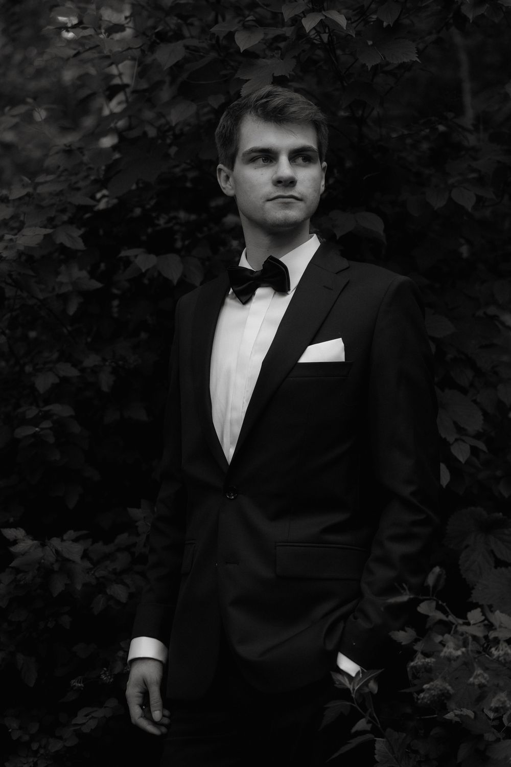 Classic black and white portrait of someone in a tuxedo with bow tie and pocket square against a leafy backdrop.