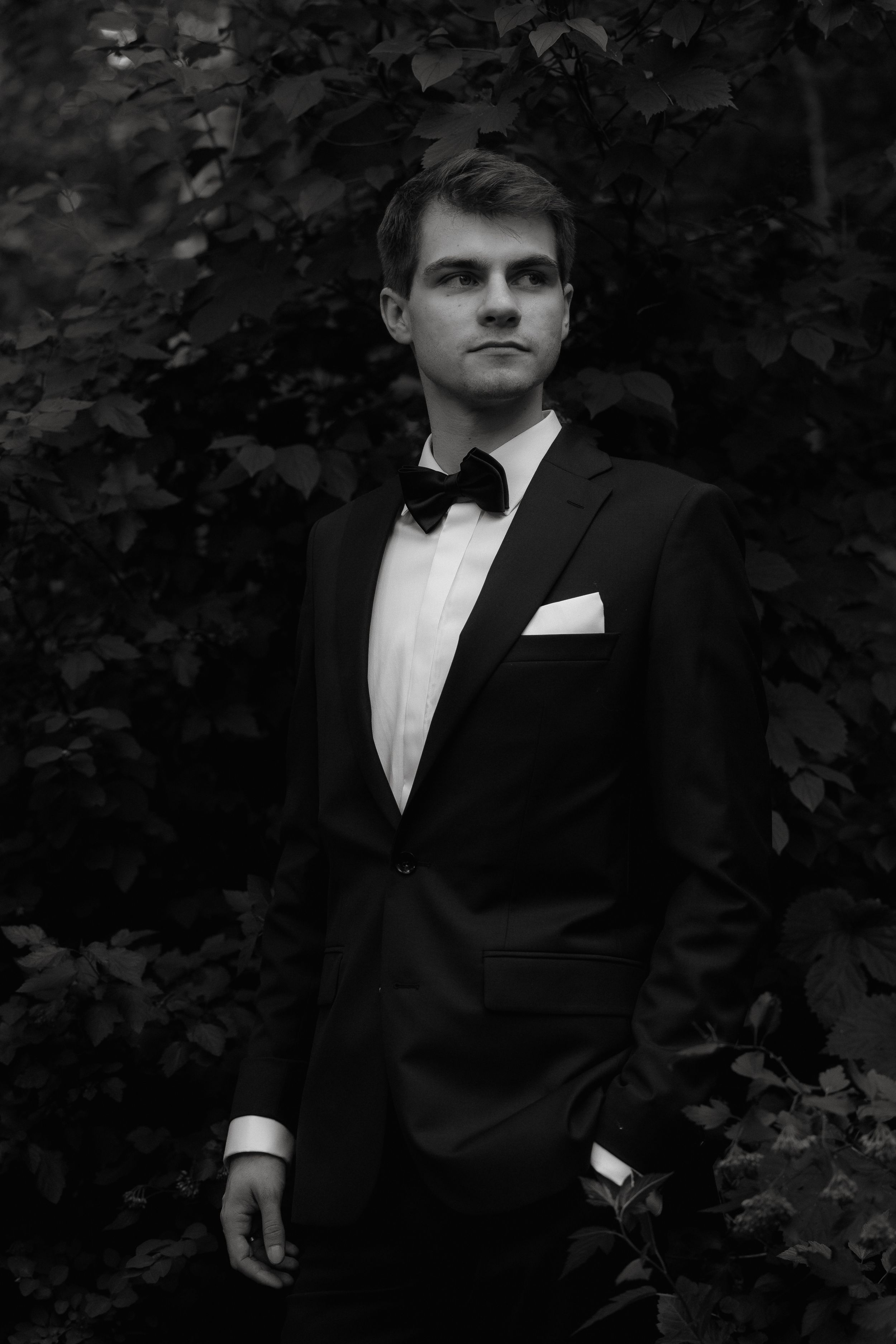 Classic black and white portrait of someone in a tuxedo with bow tie and pocket square against a leafy backdrop.