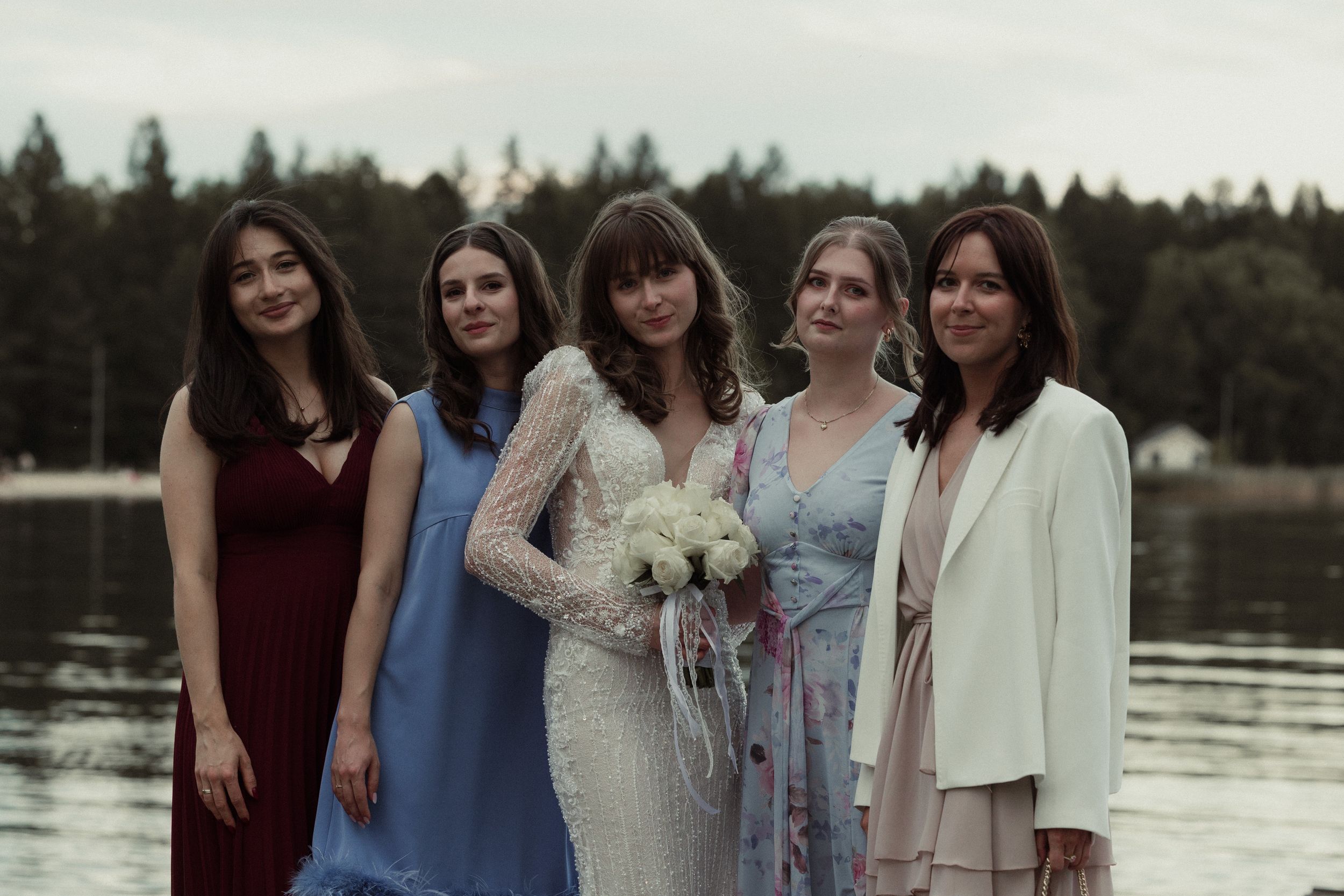 Five women stand together by a lake, with one wearing a lace wedding dress and holding a bouquet in the center.