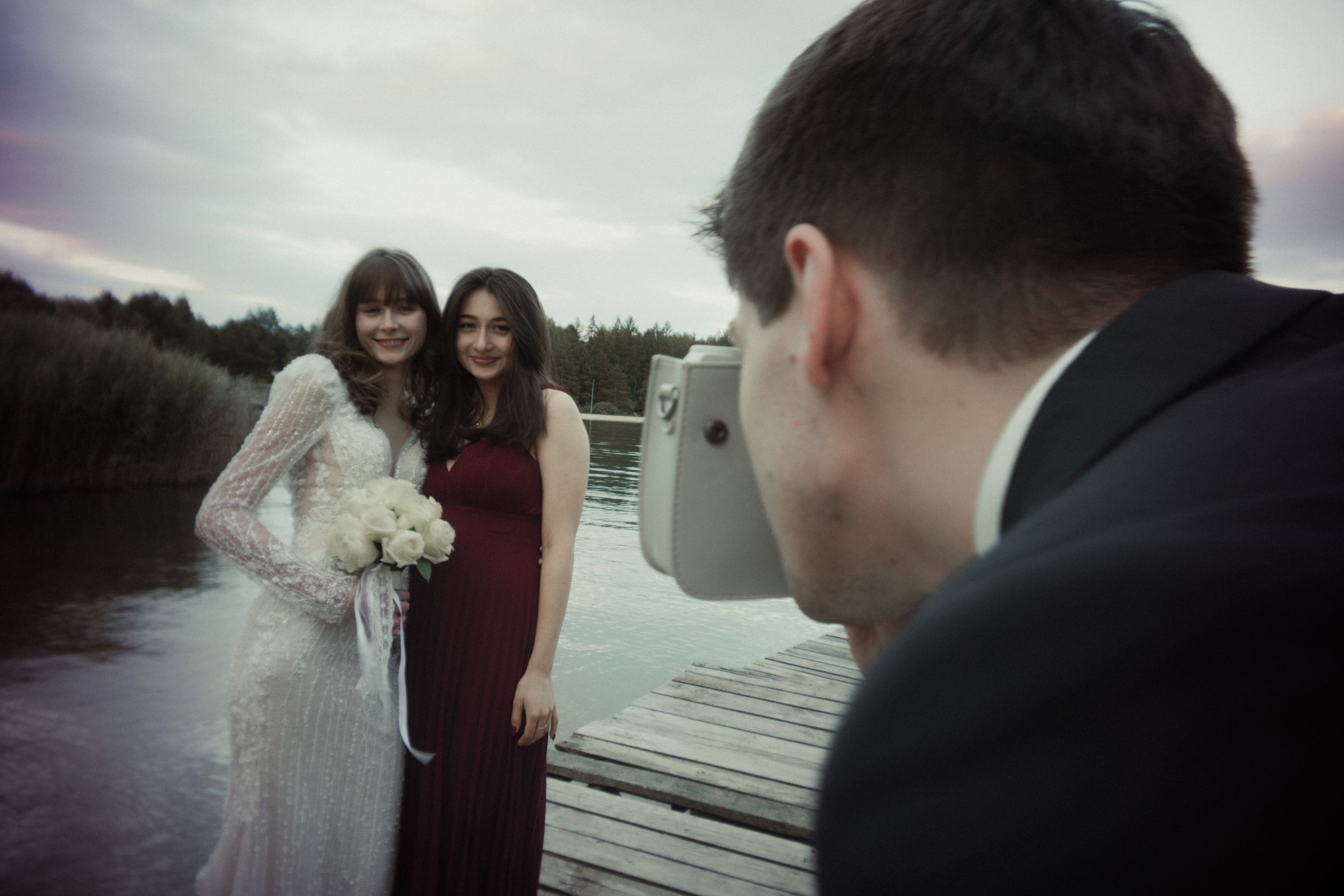 A photographer captures a wedding scene on a wooden dock by the water at dusk.