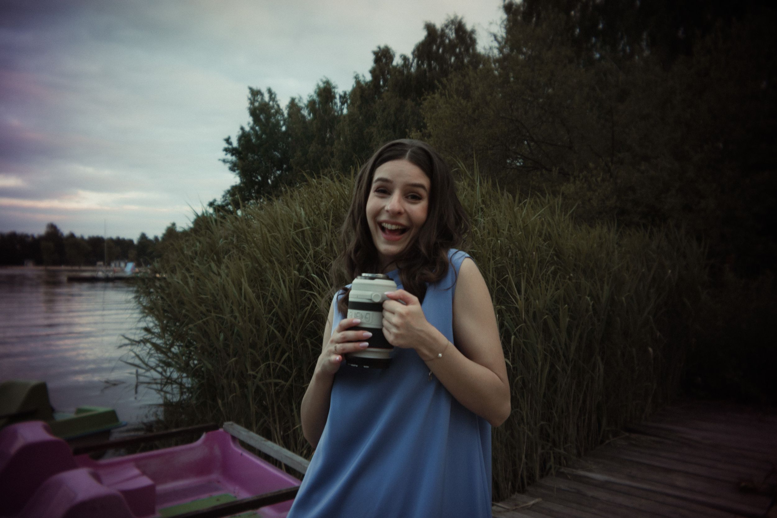 Someone in a blue sleeveless top holds a drink while standing on a dock near tall reeds at dusk beside water.
