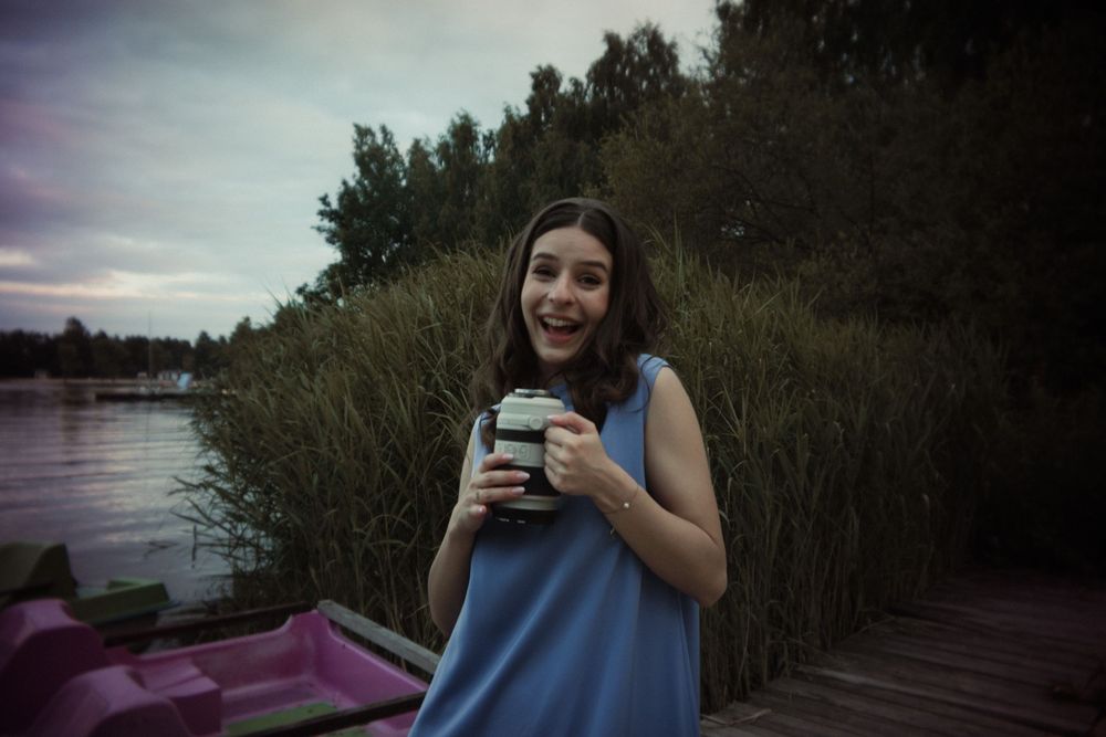 Someone in a blue sleeveless top holds a drink while standing on a dock near tall reeds at dusk beside water.