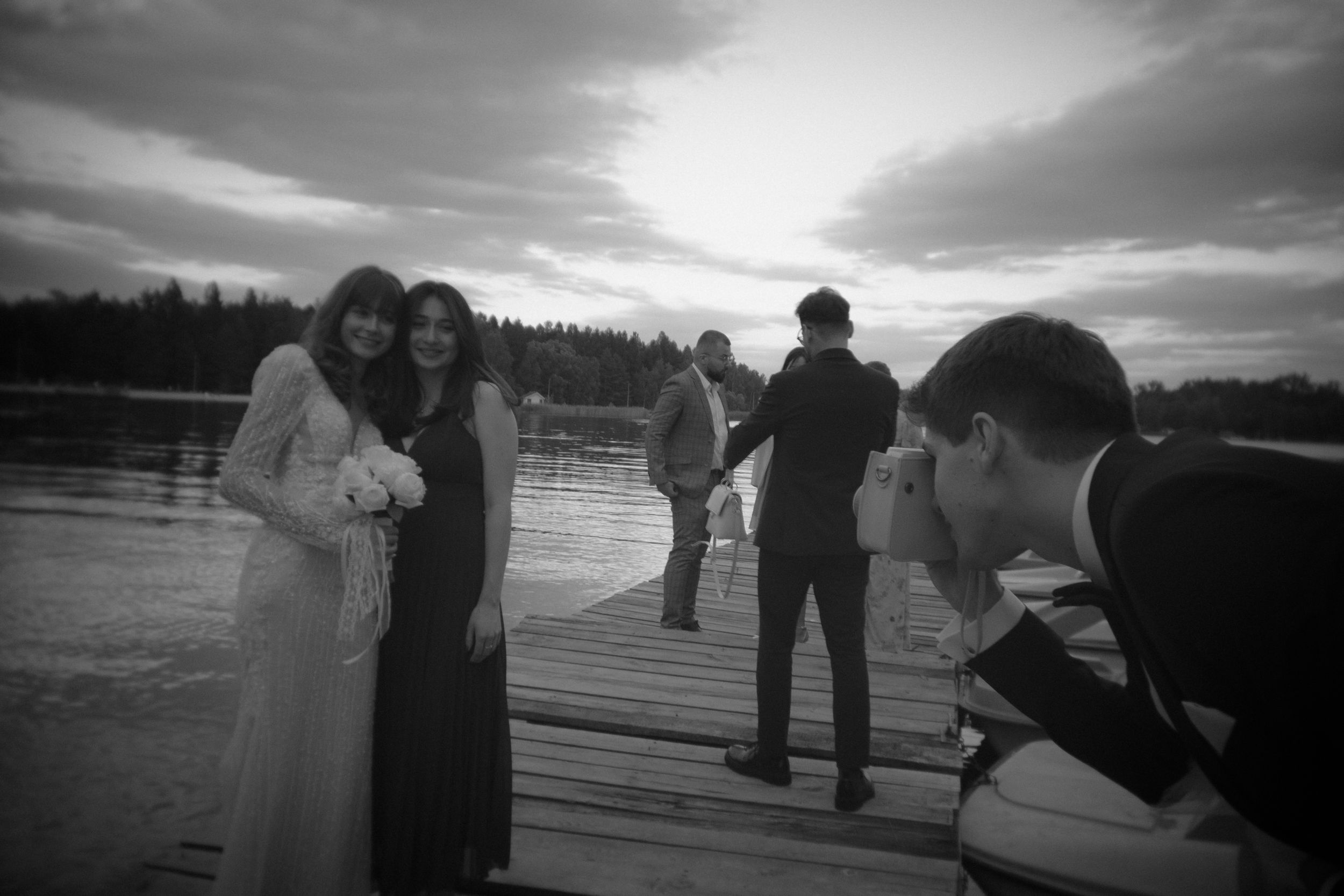 Wedding celebration on a lakeside dock with photographer capturing group photos at sunset in black and white.