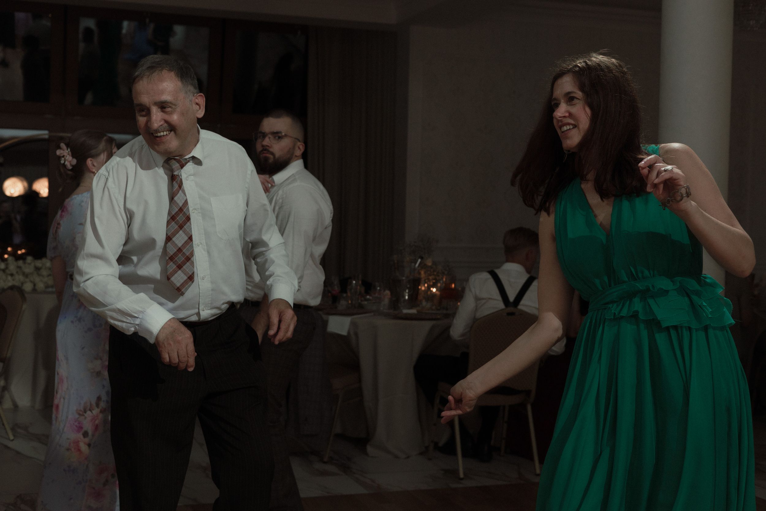Two people dancing at a dimly lit reception, with a person in a white shirt and tie and another in an emerald green dress.