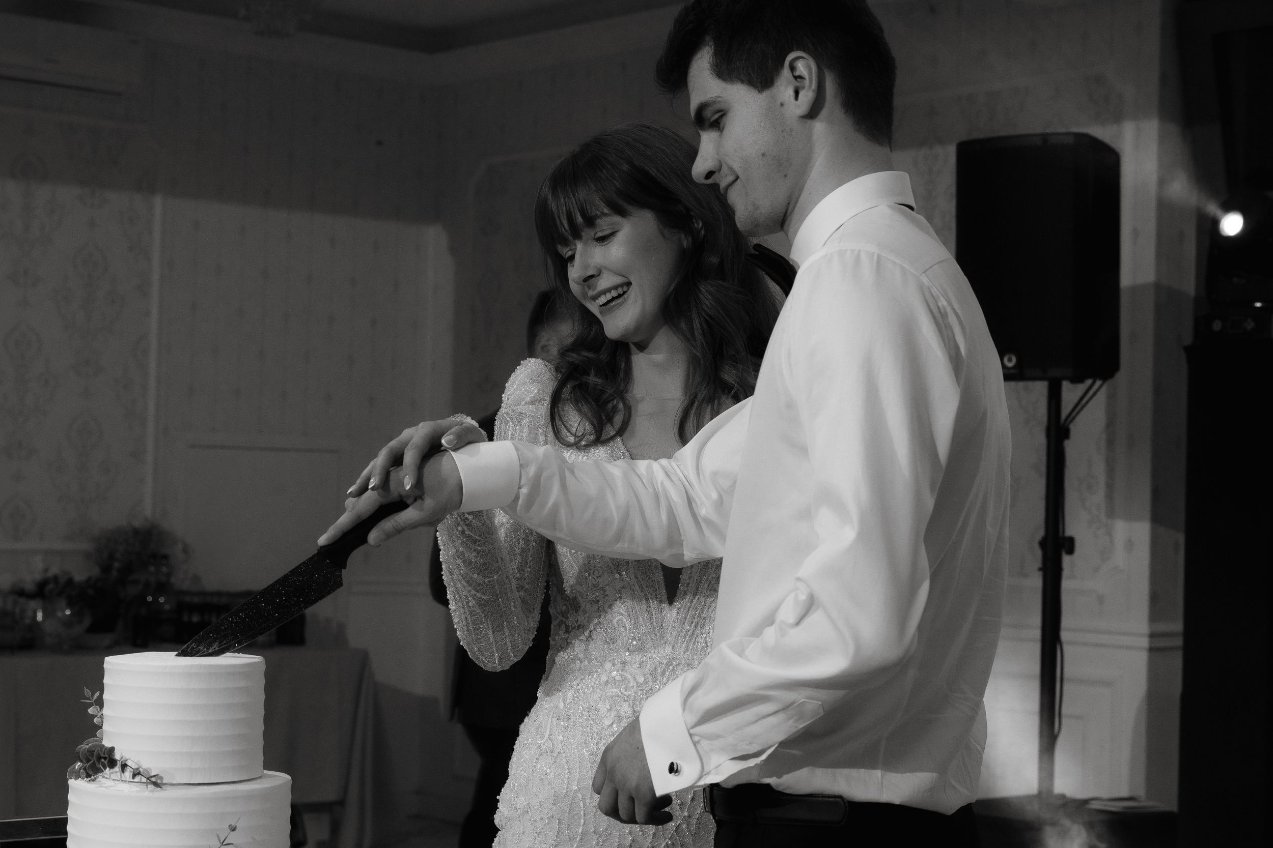 Black and white image of a couple cutting their wedding cake together at their reception celebration.