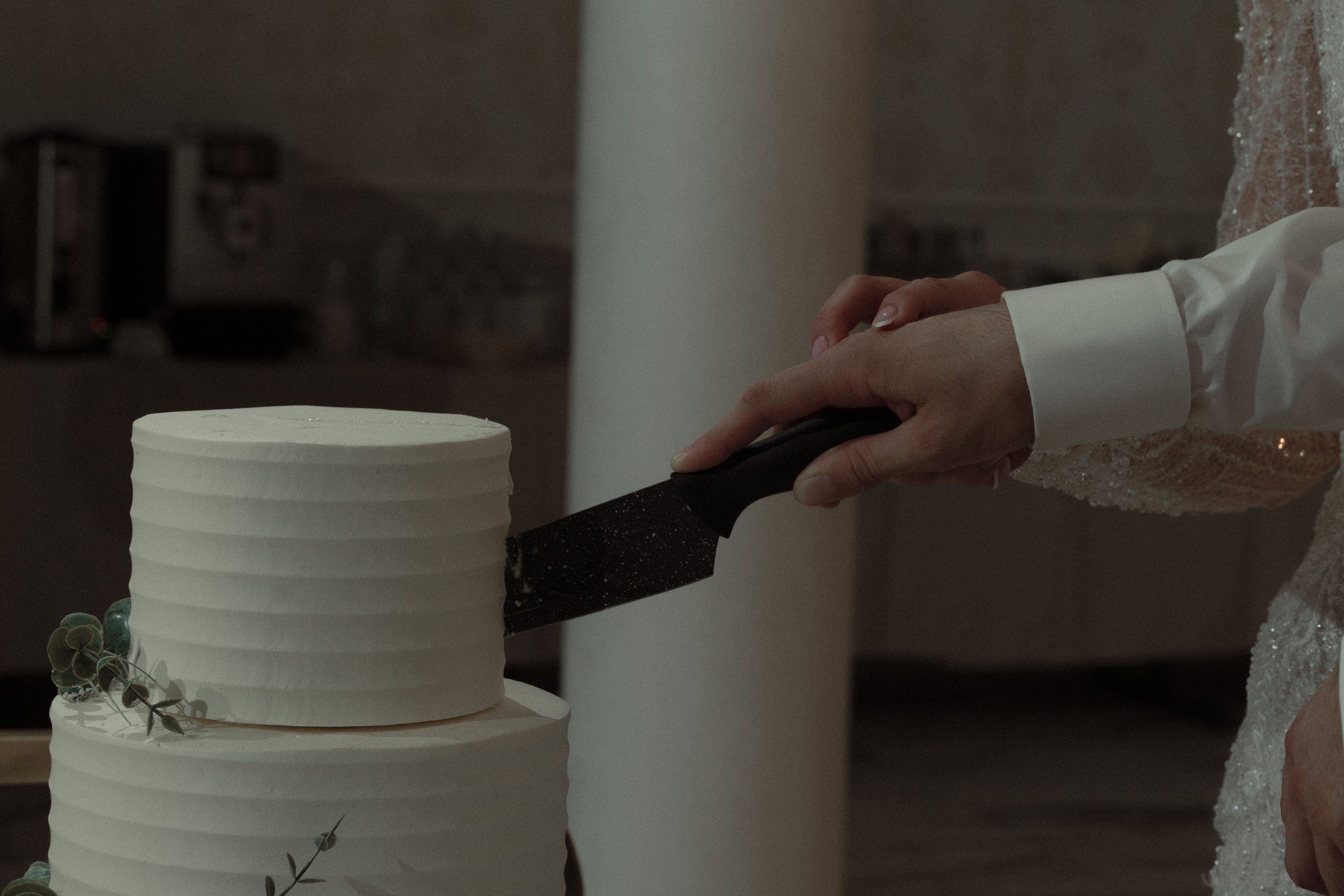 A hand holding a black icing spatula smooths the side of a two-tiered white wedding cake.