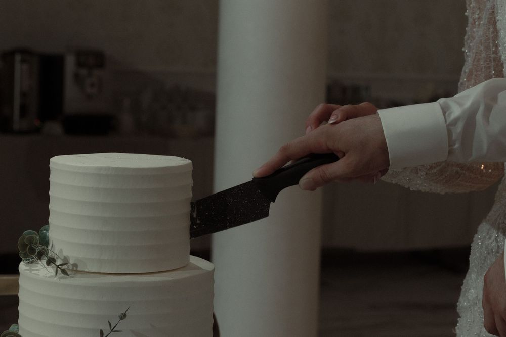 A hand holding a black icing spatula smooths the side of a two-tiered white wedding cake.