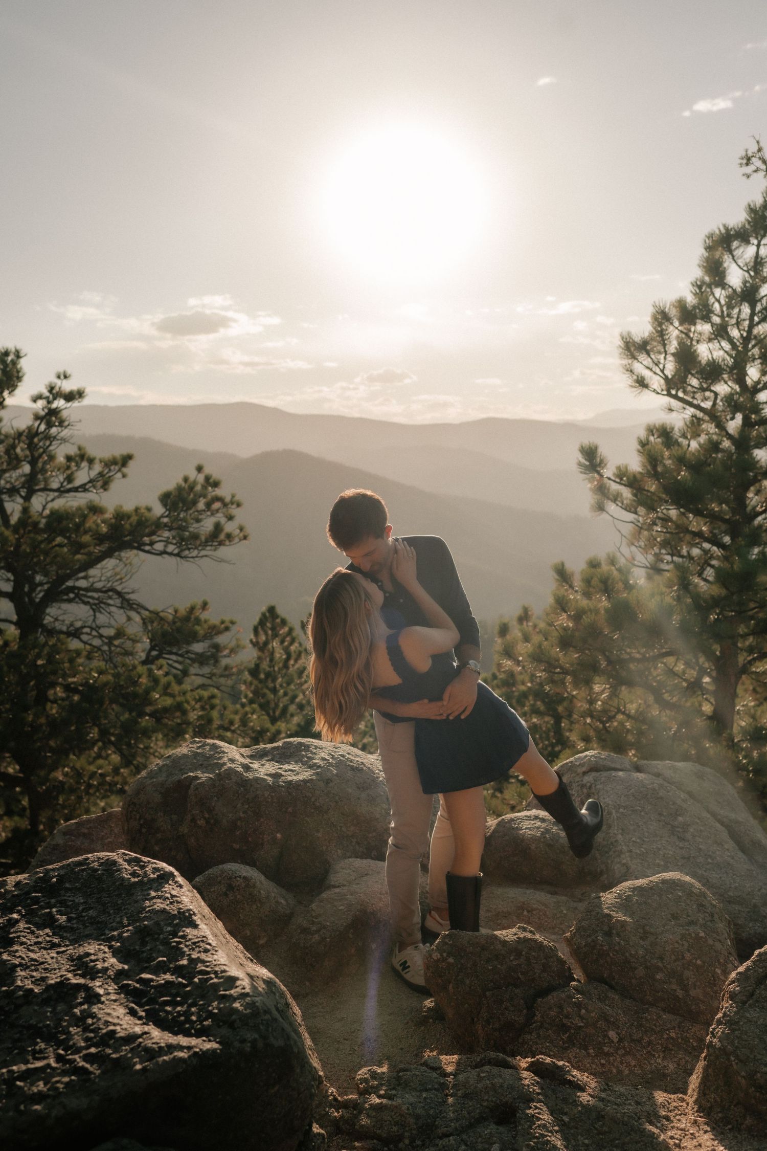 A couple embracing on a rocky mountain overlook at sunset with pine trees and distant mountain ranges in view.