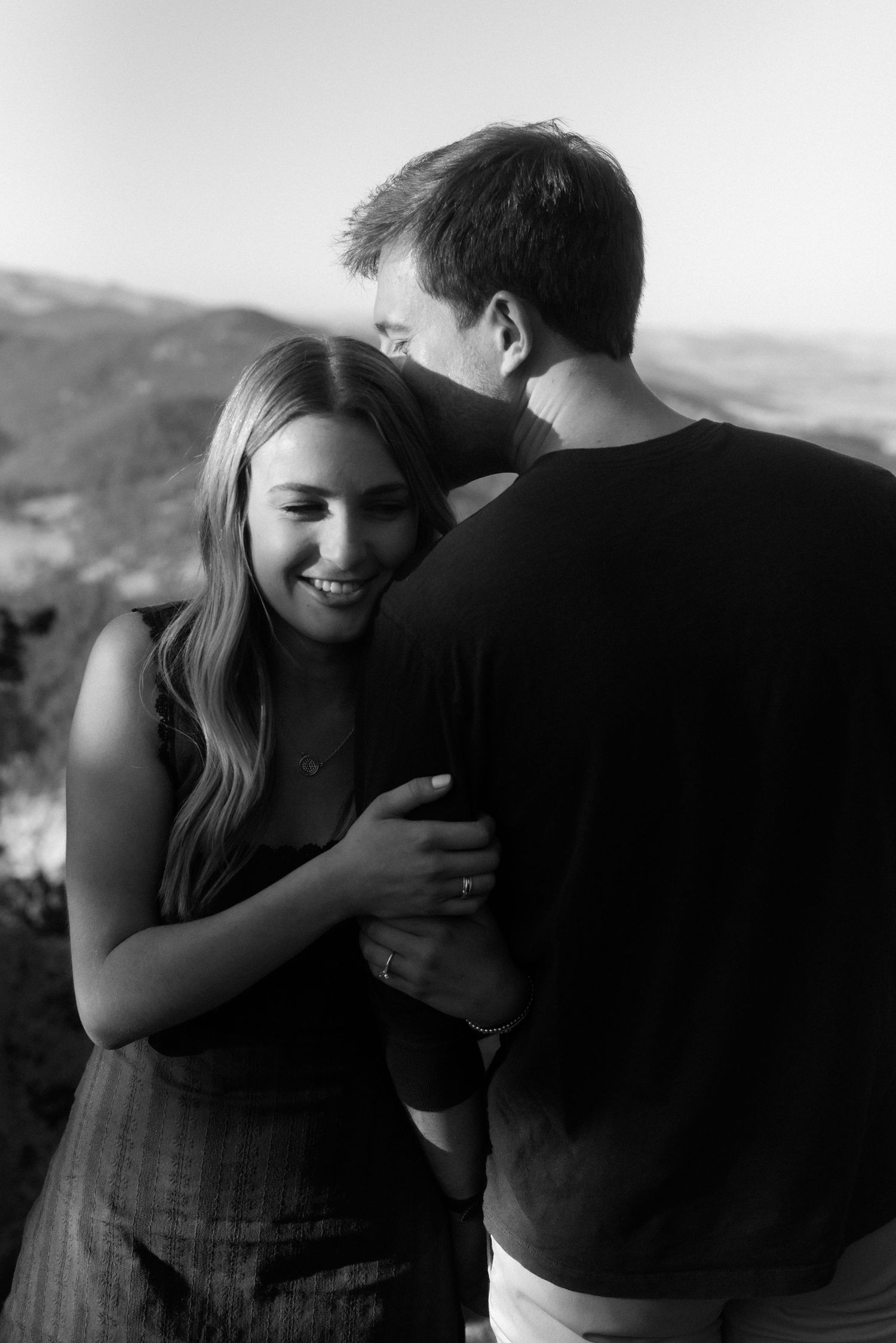 A young couple embraces outdoors in black and white, sharing an intimate moment against a scenic mountain backdrop.