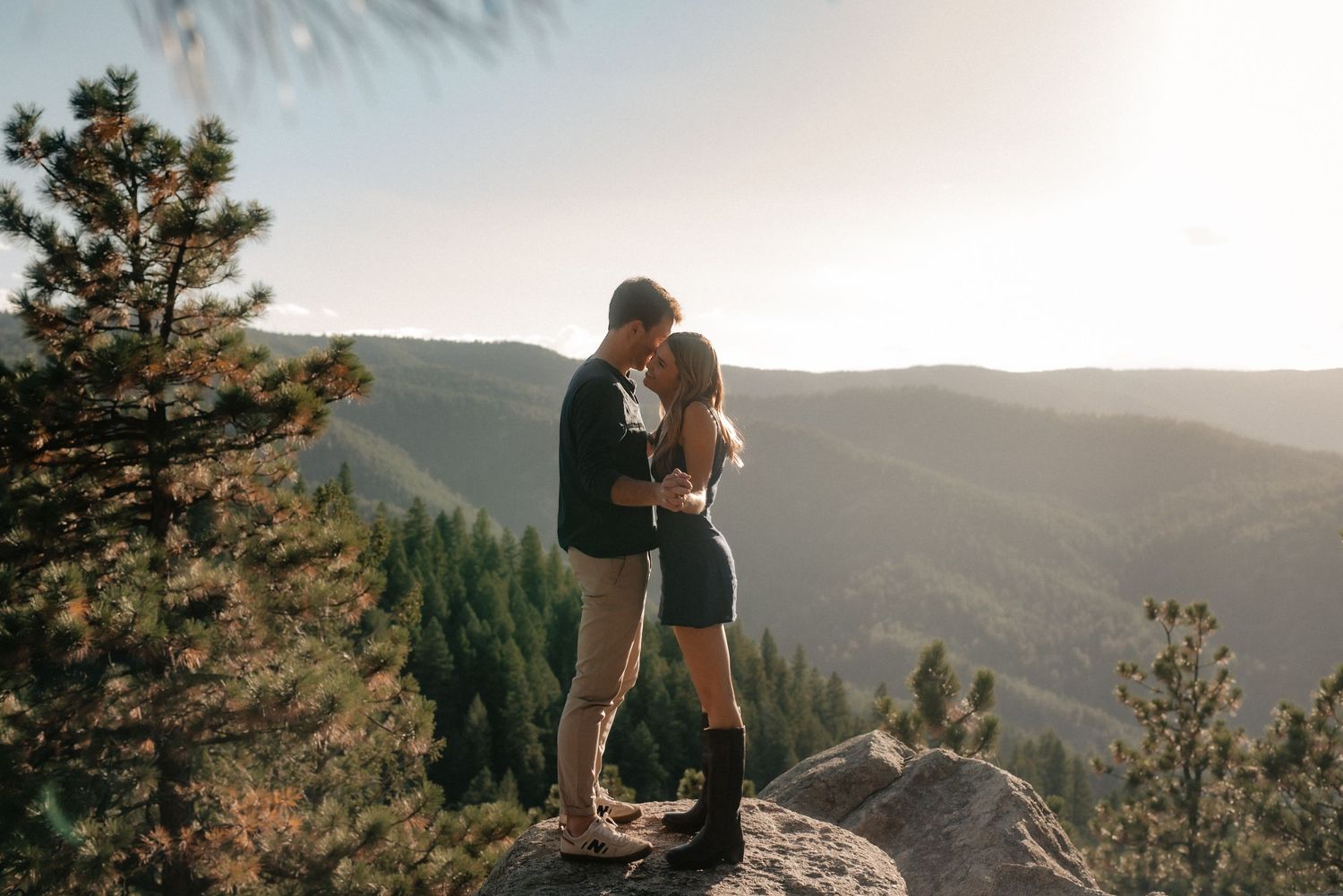 Two people share an intimate moment on a mountain overlook during golden hour with forested hills stretching into the distance.