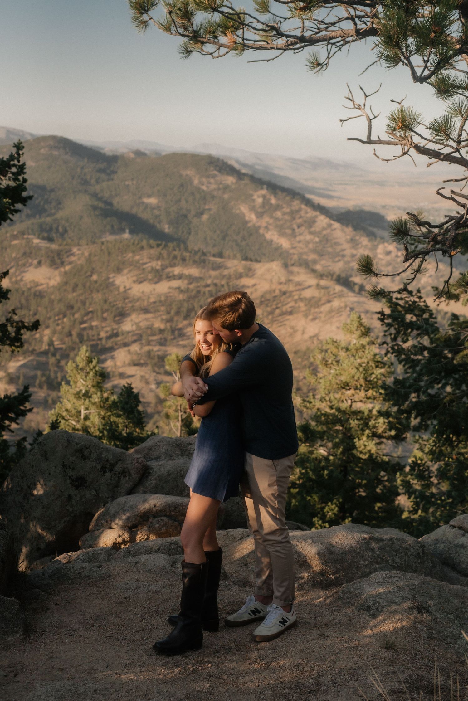 A couple embraces on a rocky mountain overlook during sunset with sweeping valley views in the background.