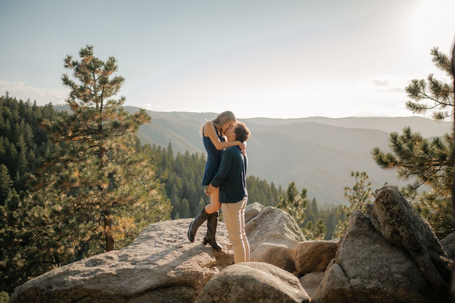 A couple embraces while standing on a rocky overlook with mountains and evergreen forest in the background.