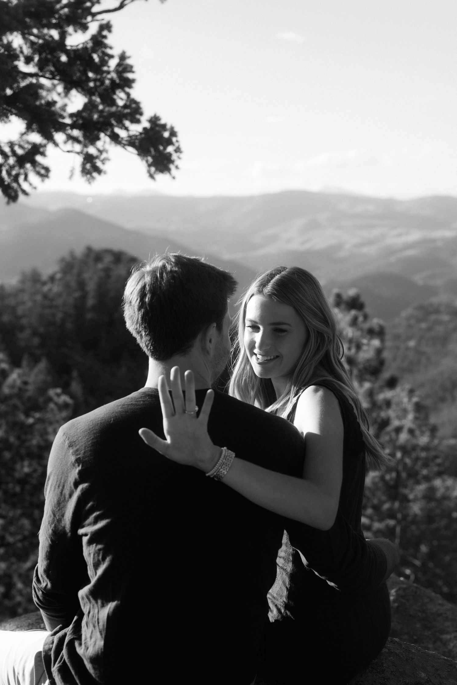 A couple sits together on a mountaintop overlooking rolling hills in black and white photography.