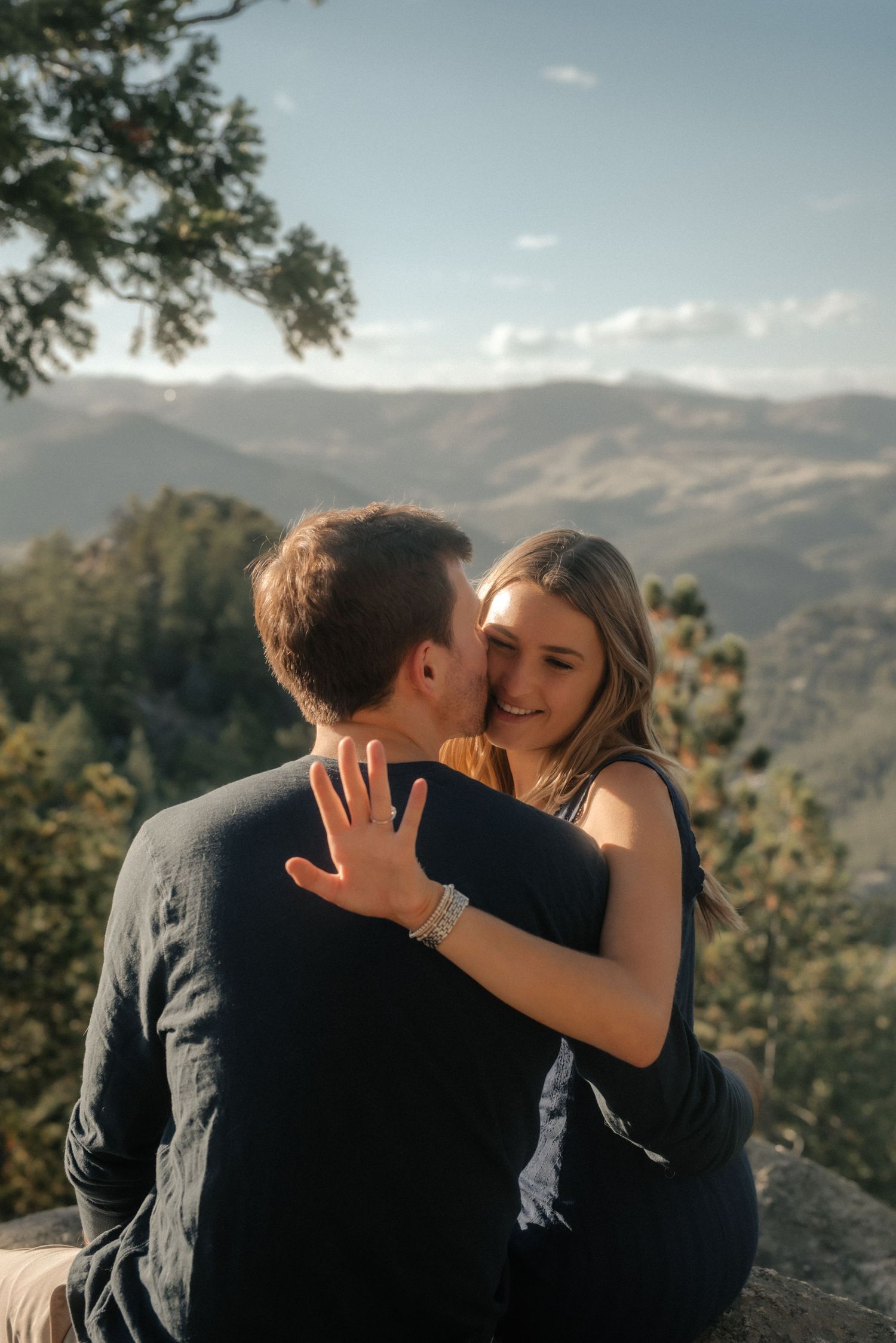 A romantic couple embraces on a mountain overlook with sweeping valley views and blue skies in the background.