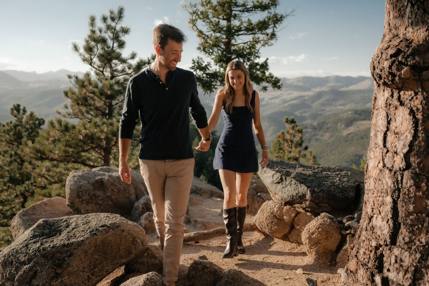 A couple walks together on a rocky mountain trail with scenic mountain vistas in the background.