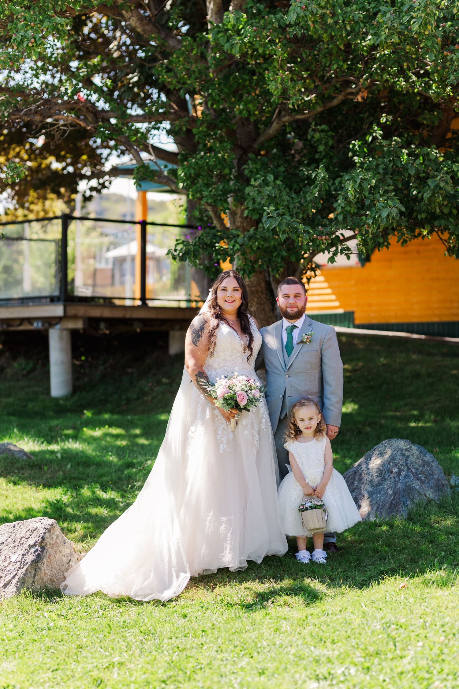 A lovely outdoor wedding scene with the couple and flower girl posed in a sunlit garden area next to a tree.