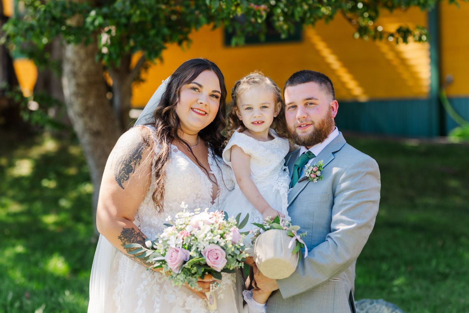 A loving family poses together at an outdoor wedding with floral bouquet on a sunny day against a yellow wall.