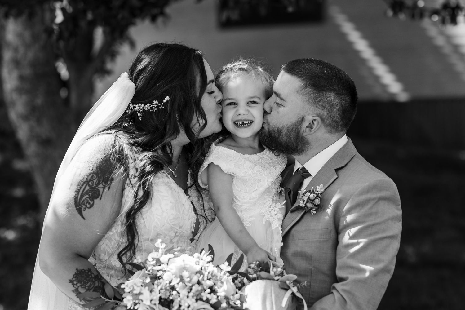 A black and white wedding portrait showing a couple in wedding attire sharing a tender moment with a young girl in white dress.