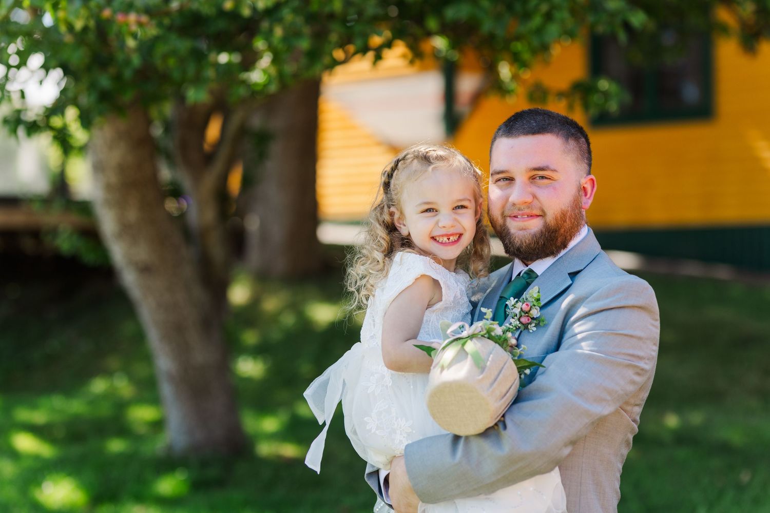 A tender moment captured outdoors with a small child in a white dress and an adult in a gray suit during a special occasion.
