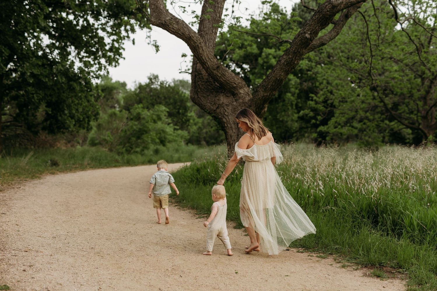 A mom and her toddlers walk hand in hand through an outdoor path surrounded by greenery and natural lighting. san marcos, texas photographer