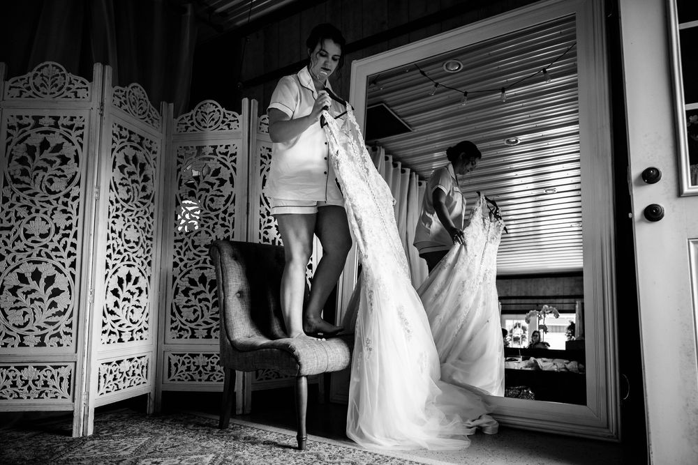 Person standing on chair helping with wedding dress preparations.