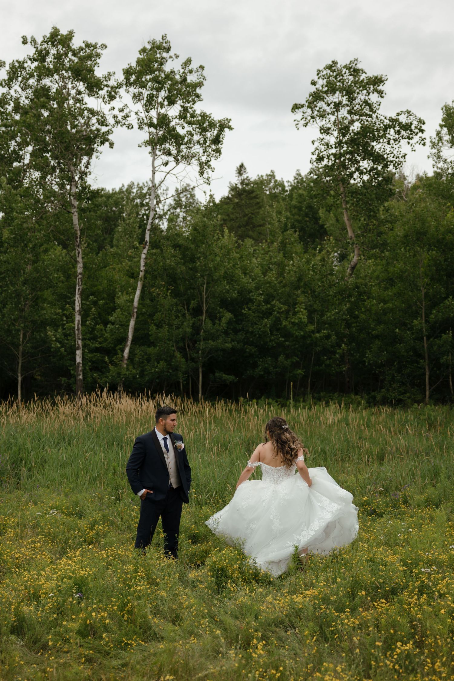 A wedding couple walks through a lush field of yellow wildflowers with birch trees in the background.