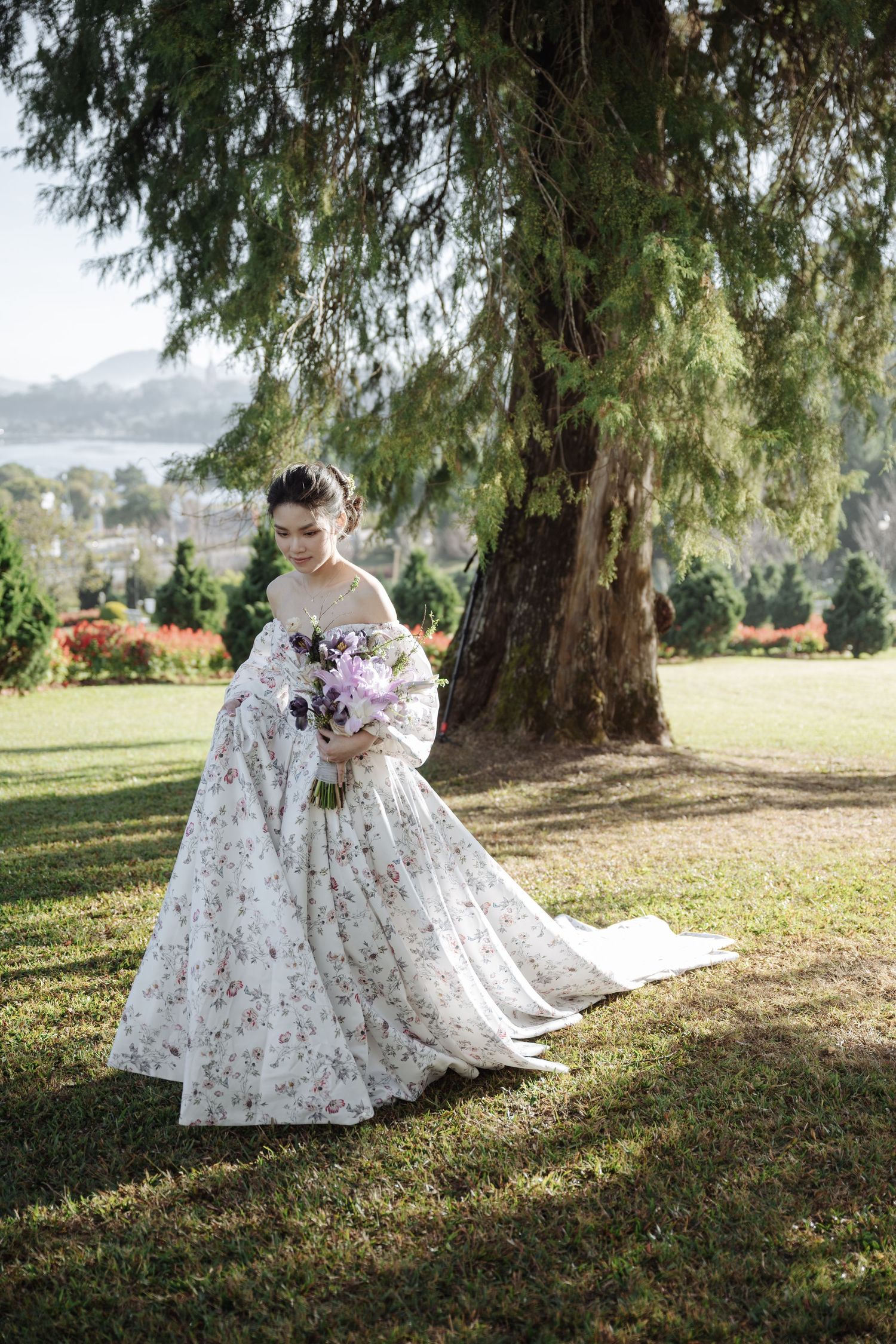 Elegant bride in floral off-shoulder ball gown holding purple bouquet beneath large tree in sunlit garden.