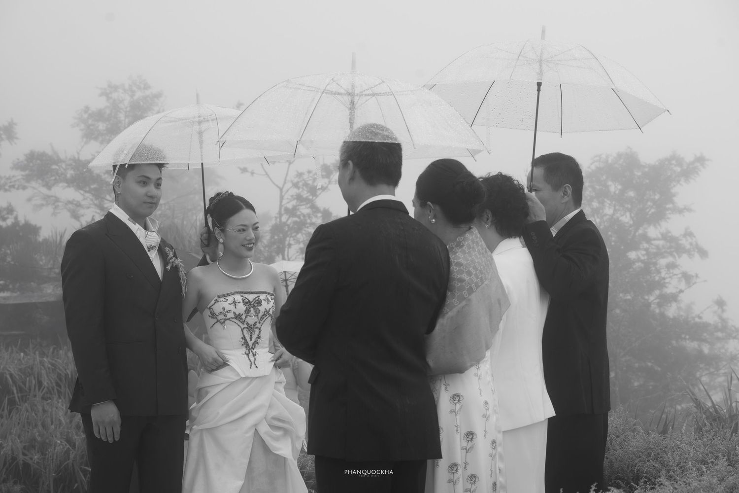 Wedding group gathers under umbrellas in foggy outdoor setting, captured in classic black and white.