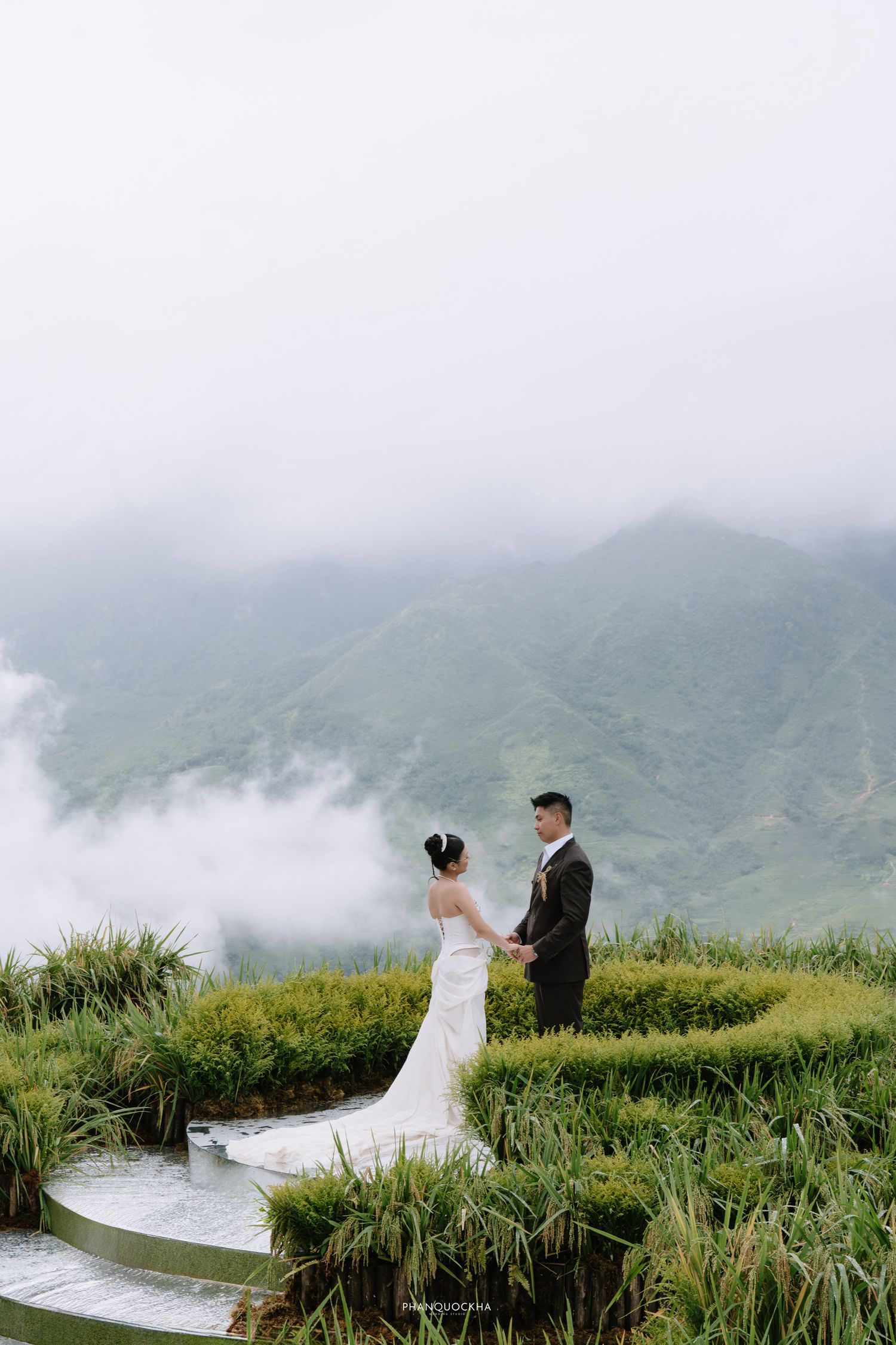 Wedding couple holds hands on lush green terraced hillside with dramatic cloudy mountain views.