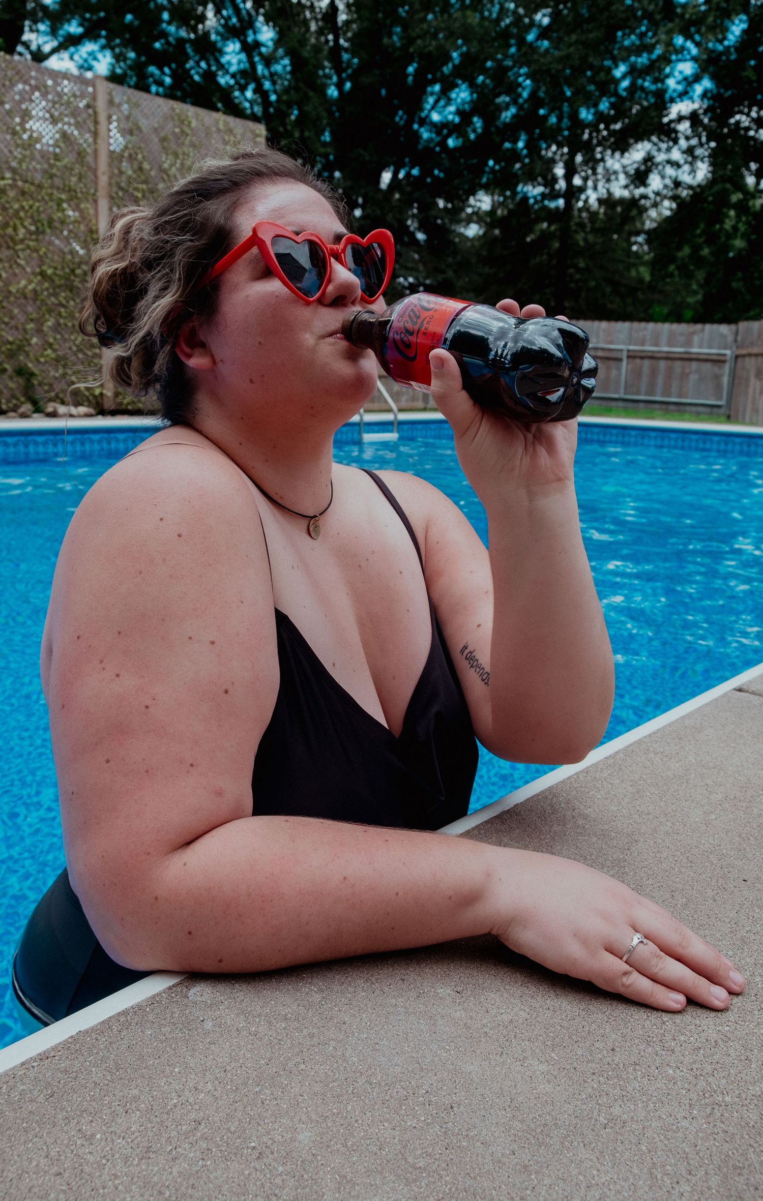 Someone in heart-shaped sunglasses drinks a Coca-Cola while relaxing at the edge of a swimming pool on a sunny day.