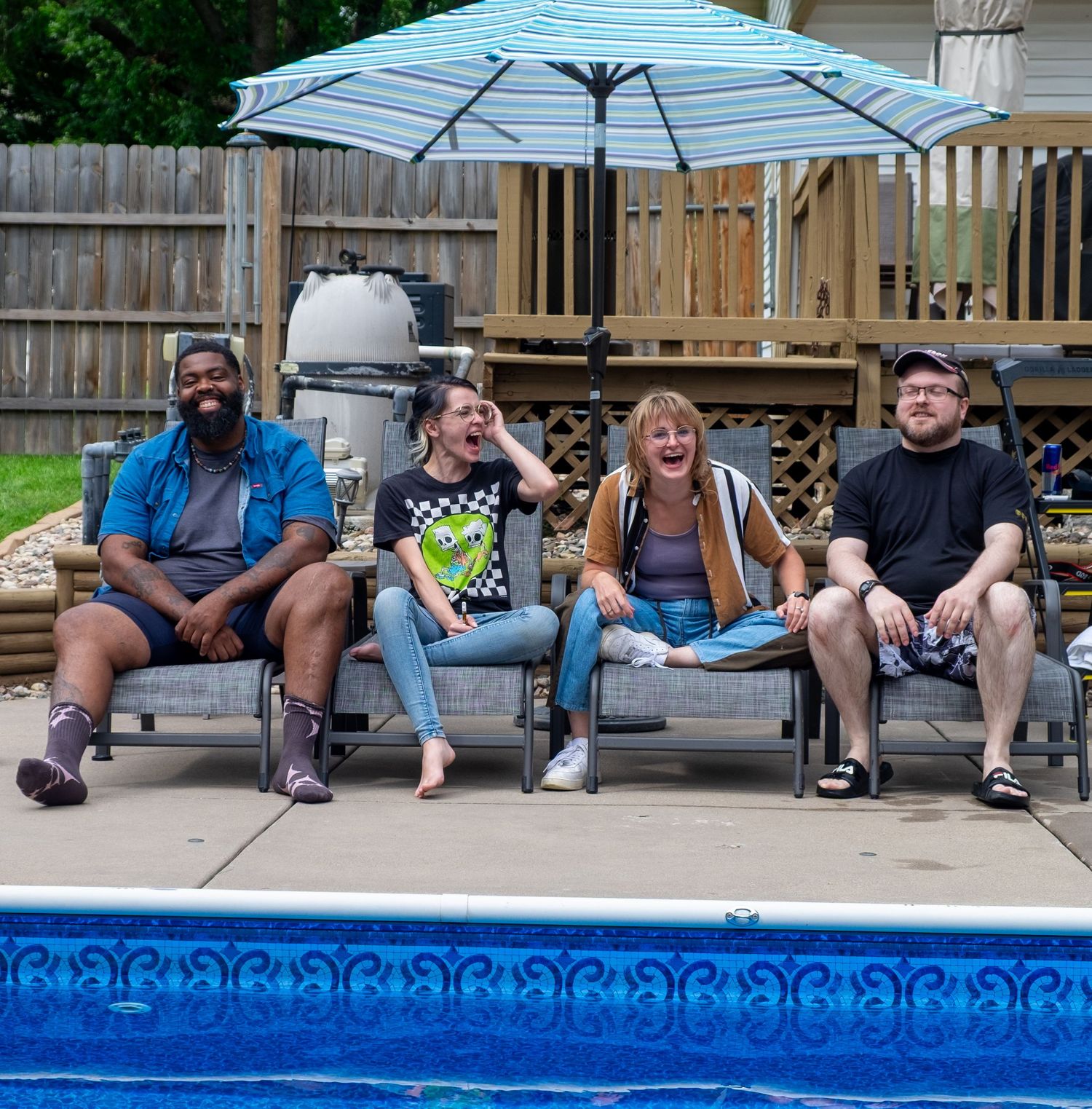 A group sits together on a poolside deck under a blue striped umbrella on a summer afternoon.