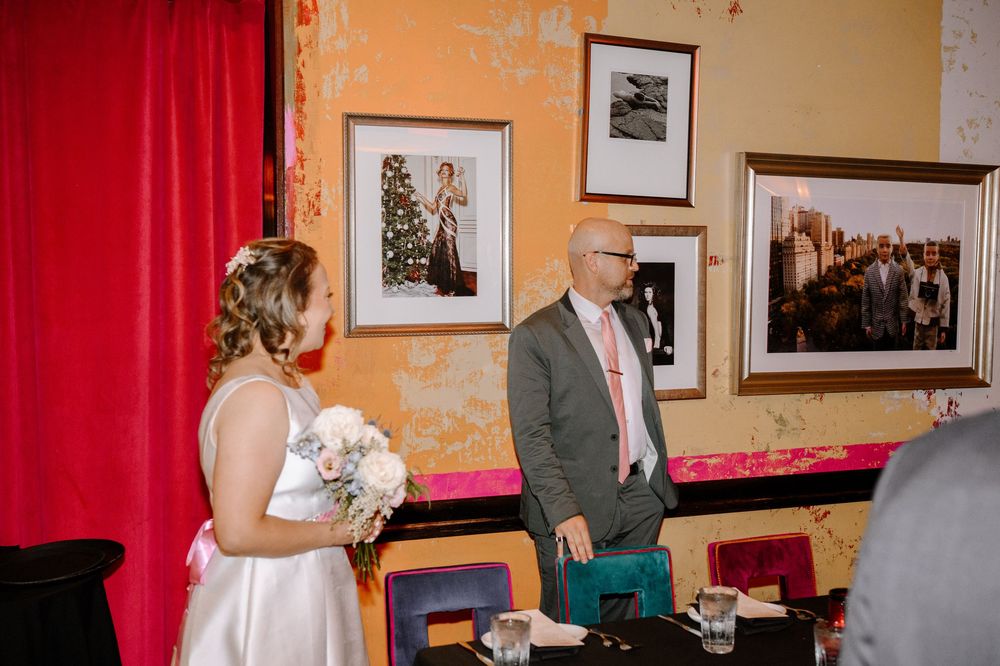 A wedding ceremony takes place in front of vibrant red curtains with framed photos on display along the walls.