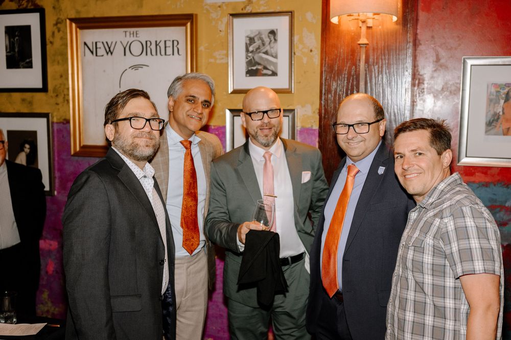 Four people converse at an event in business attire with framed artwork visible on red walls in the background.