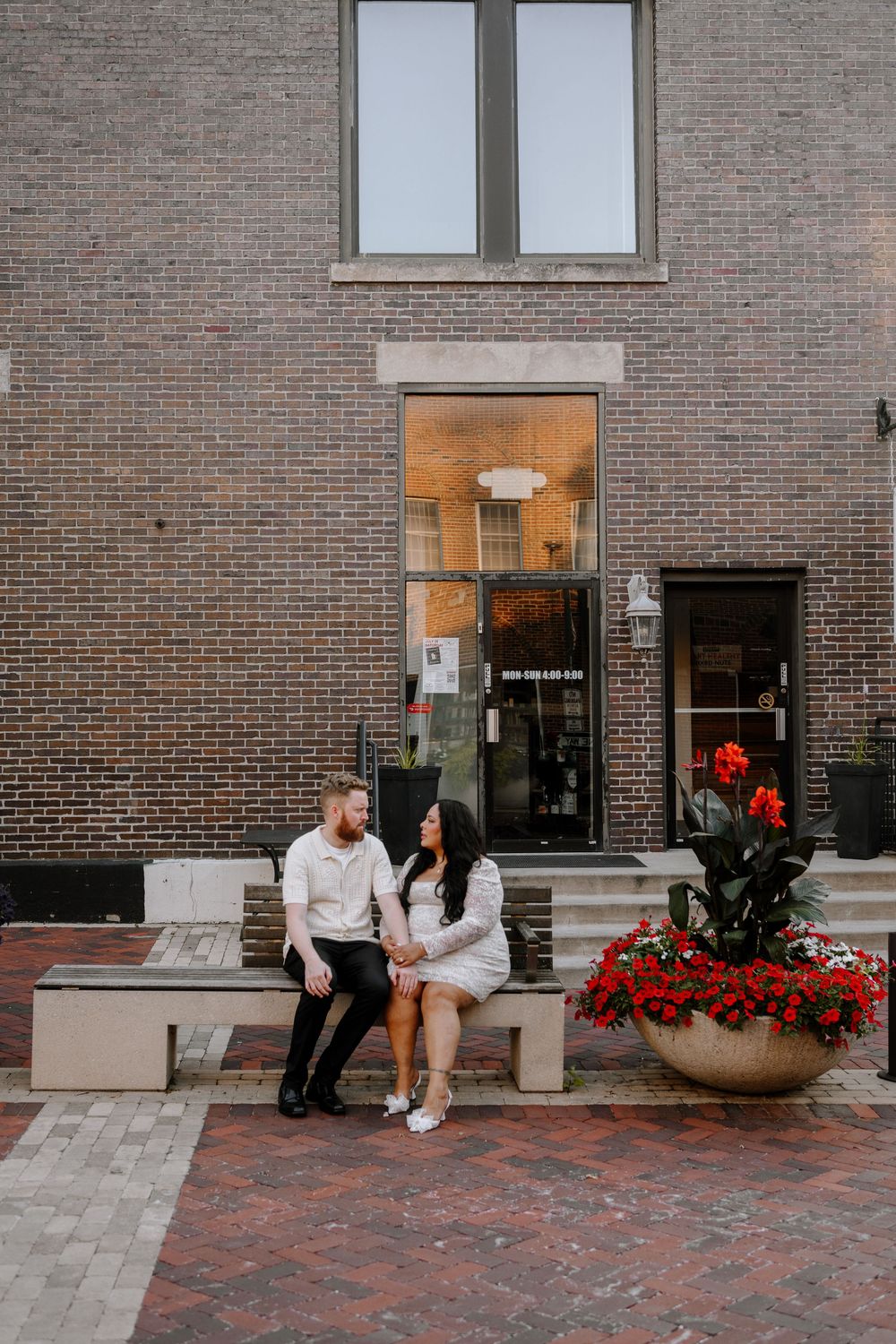 Romantic couple sitting together on brick steps with red flowers in an urban setting.