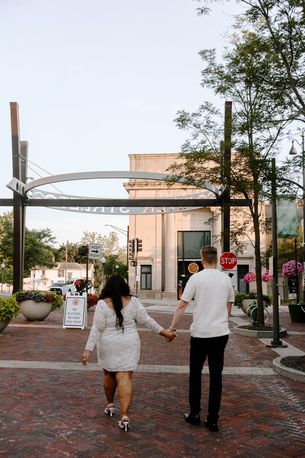 Two people walk along a brick-paved urban street with storefronts in the background.
