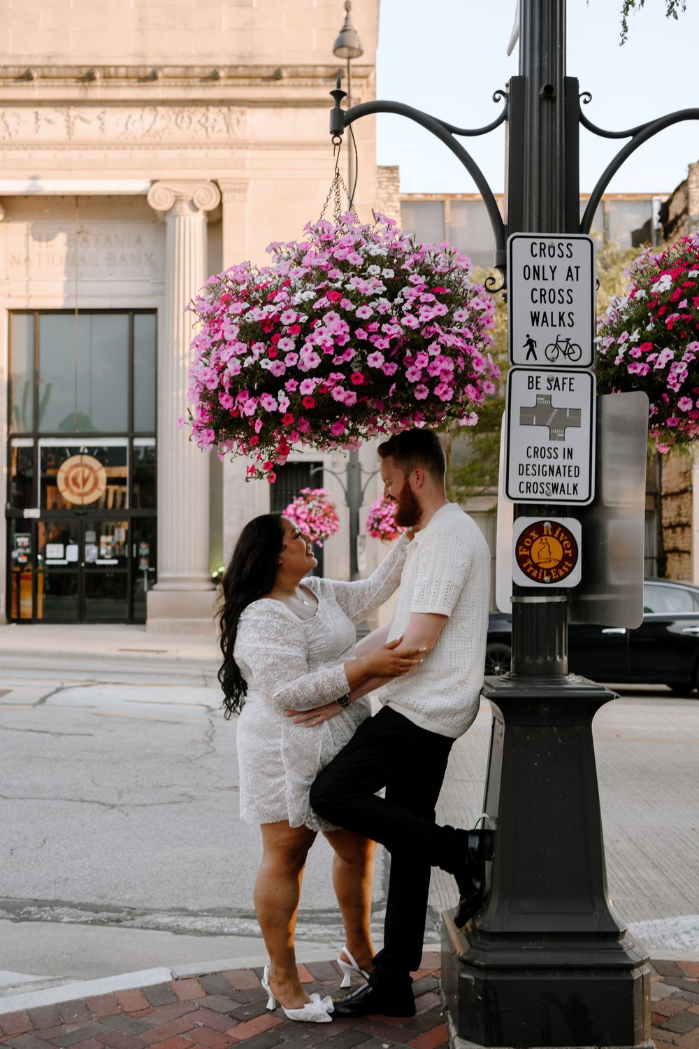A romantic moment beneath hanging flower baskets on a city street with decorative lampposts.