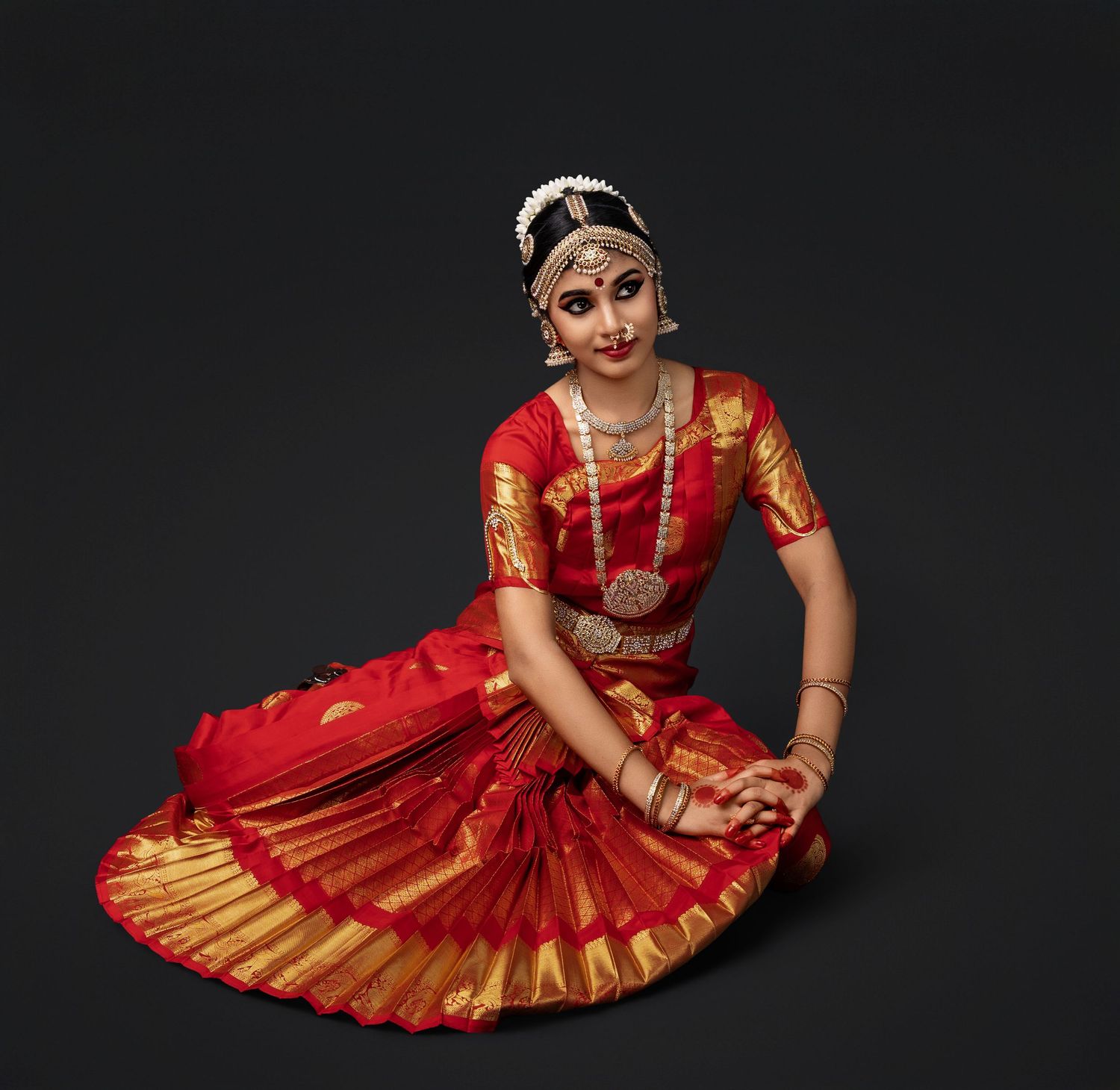 Traditional dancer in red and gold Bharatanatyam costume performs graceful poses against dark backdrop.