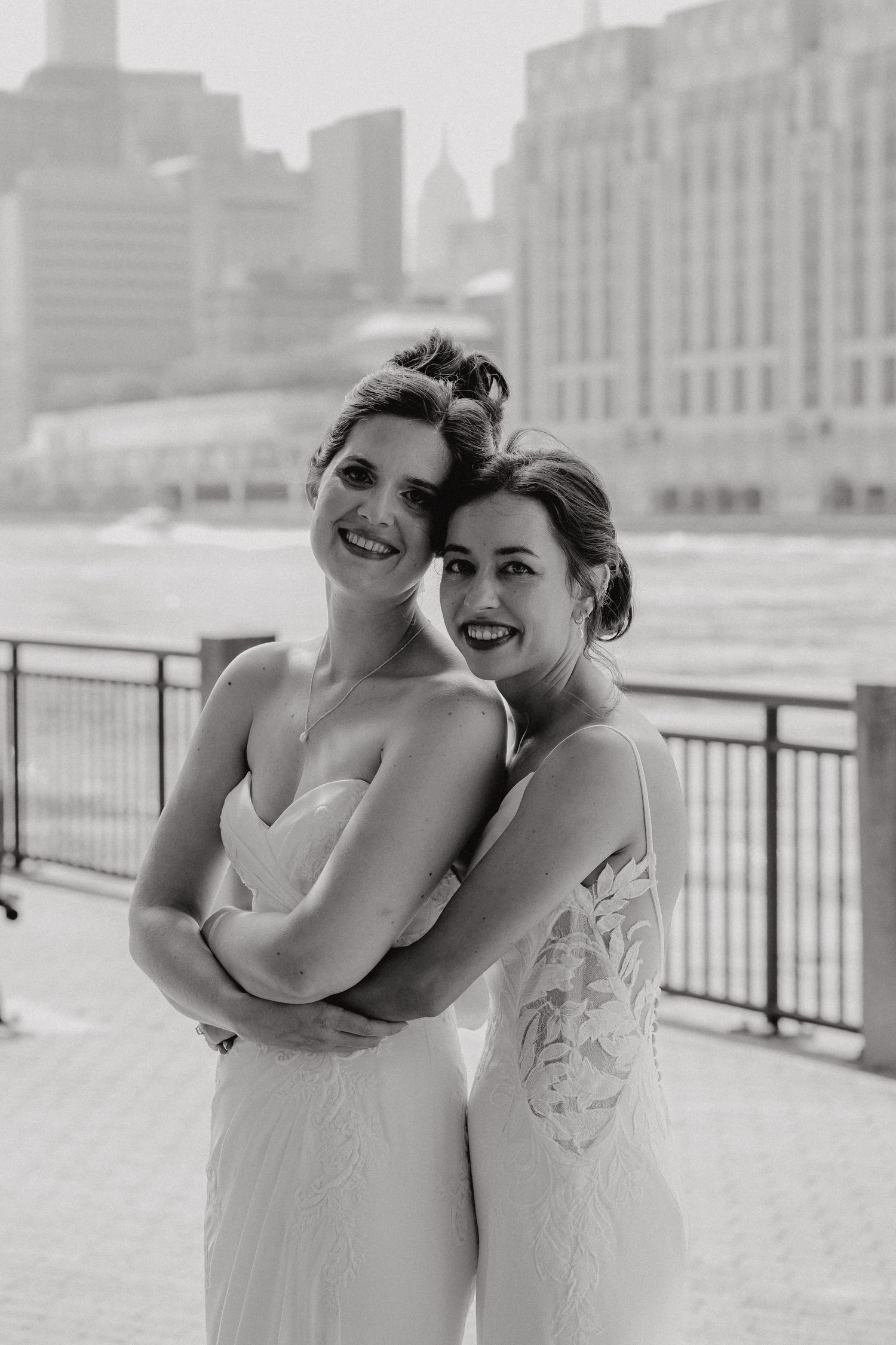 Two people in white dresses laughing together against a cityscape backdrop on a waterfront promenade at the Sanctuary Roosevelt Island NYC