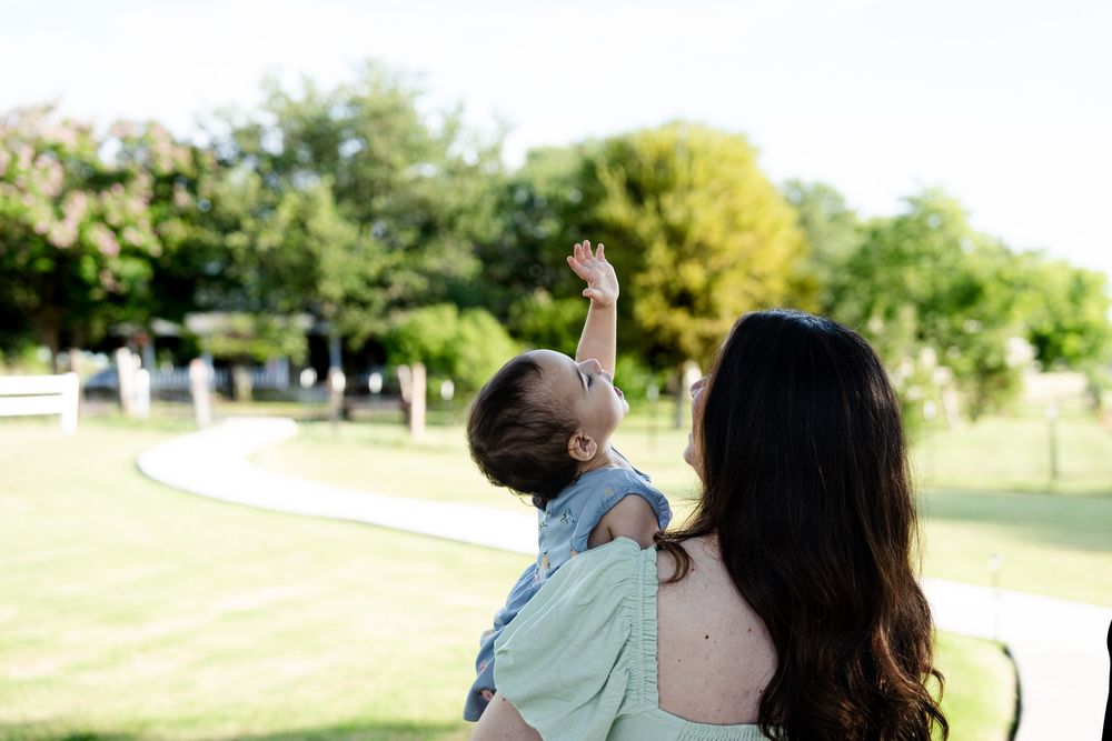 Young child points at something in the distance while being held in the park on a sunny day.