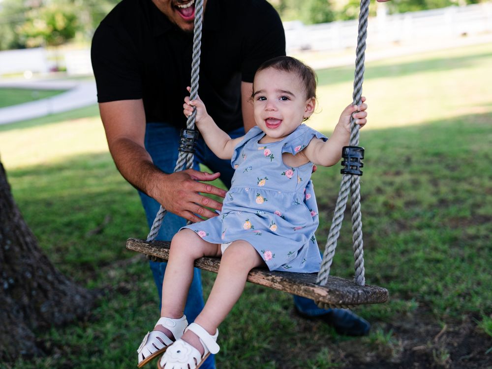 A toddler wearing blue overalls enjoys a fun moment on a swing in a sunny outdoor park setting.