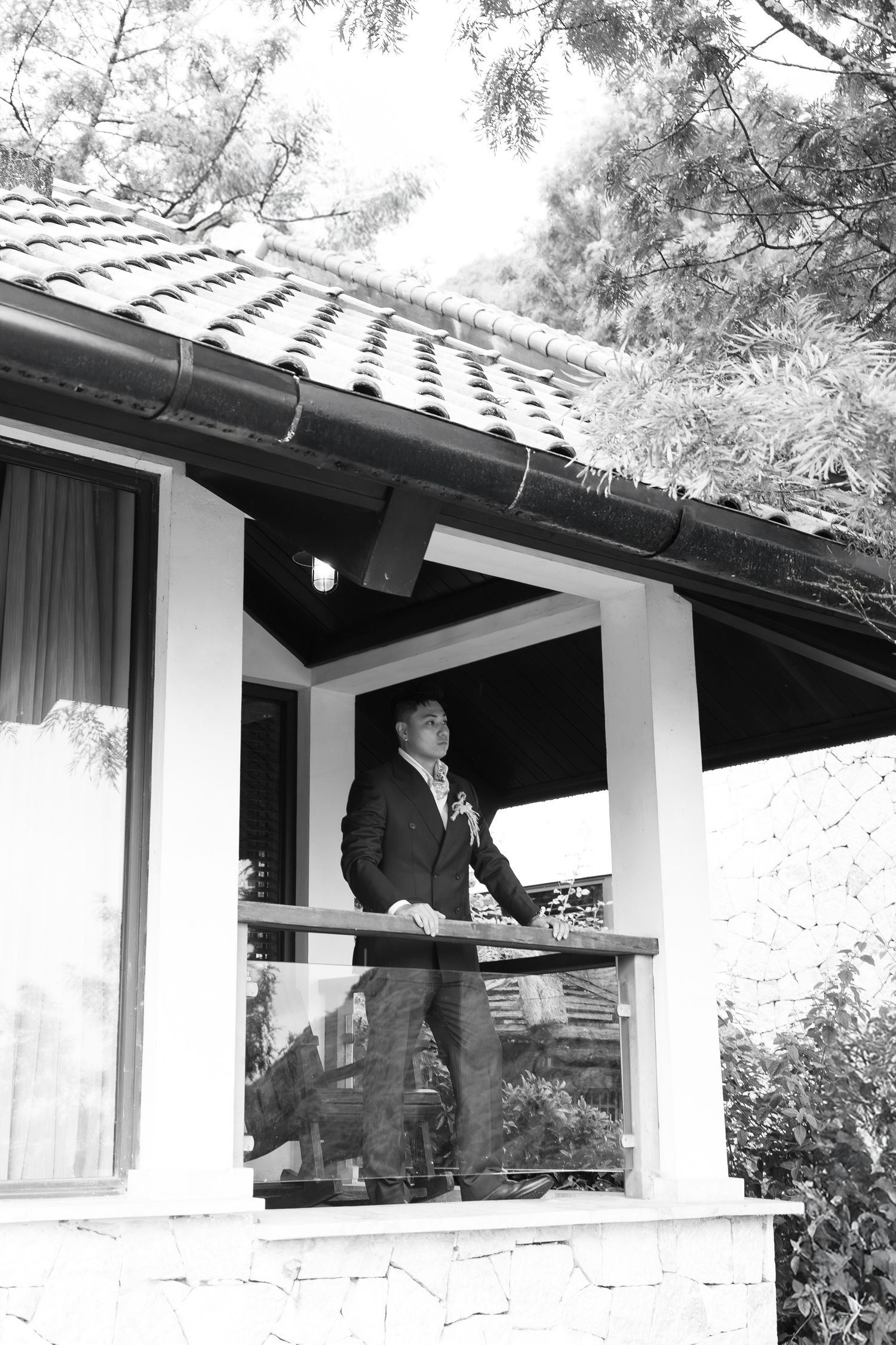 Black and white photo of groom in suit standing on a villa balcony surrounded by pine trees.