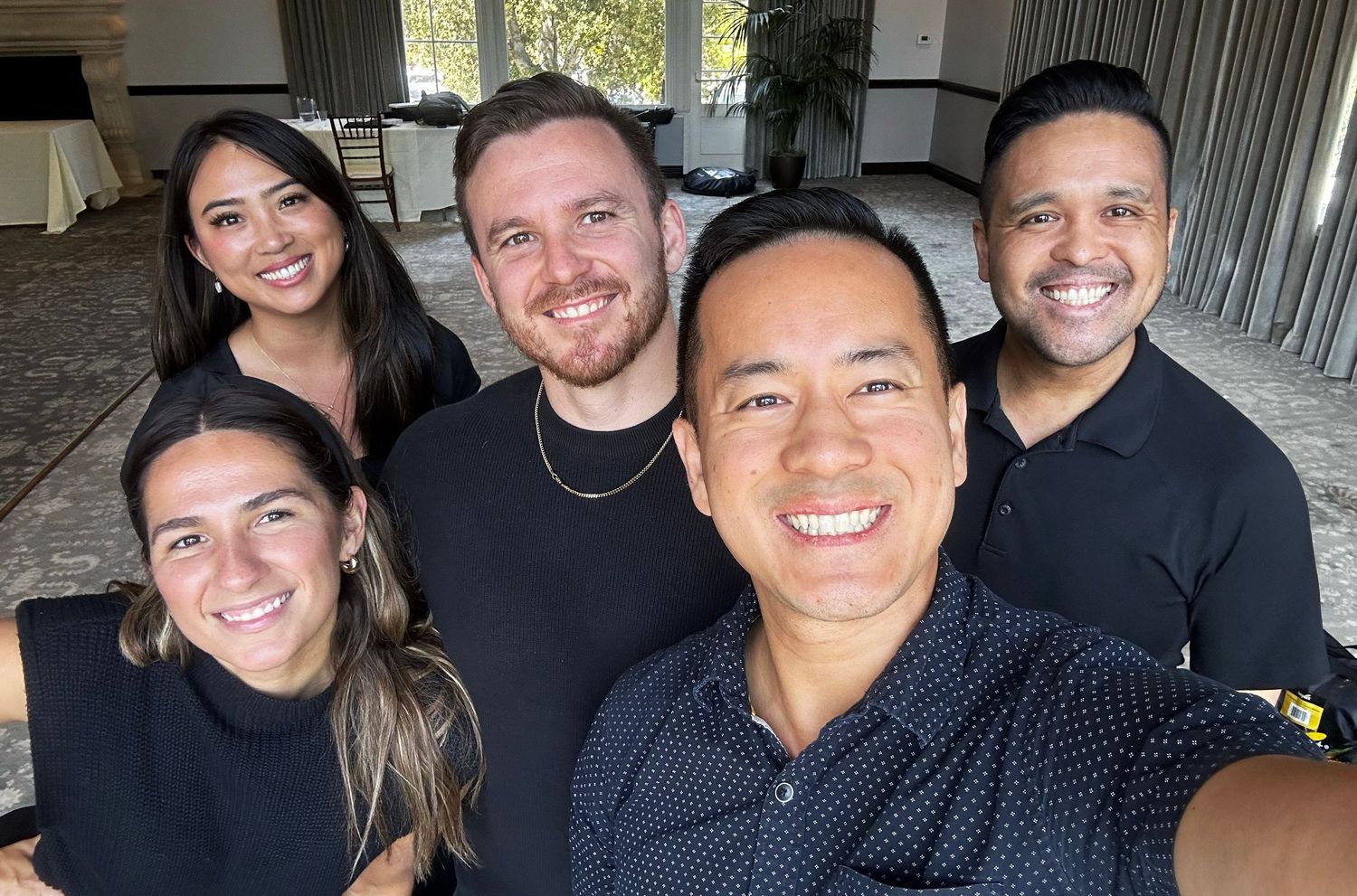 A group of five smiling wedding professionals take a selfie together in a bright event venue.