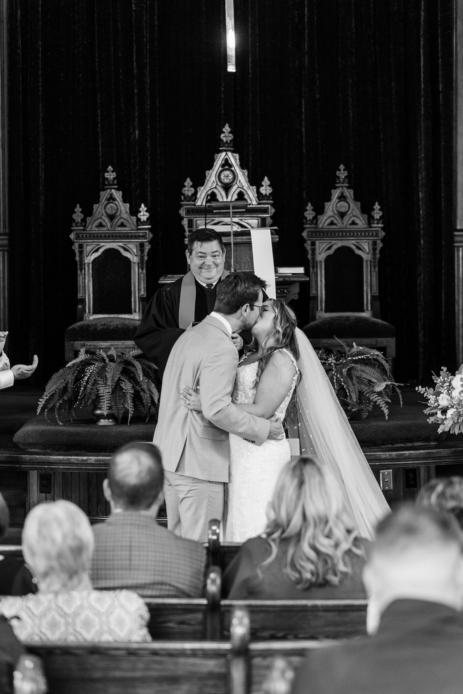 A black and white wedding ceremony photo shows a couple at the altar with ornate gothic church chairs in the background.