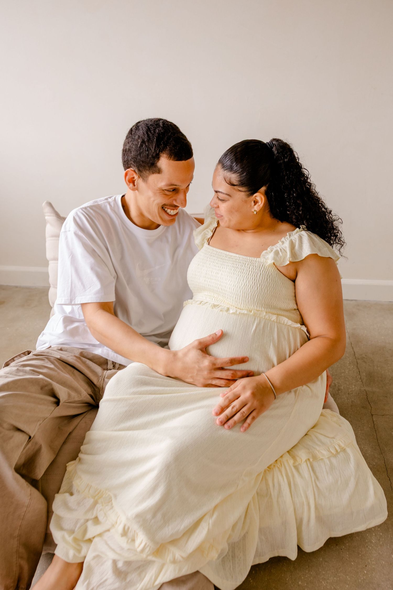 Romantic pregnancy photoshoot with couple in matching white outfits lounging together on grey sofa sharing intimate moments.
