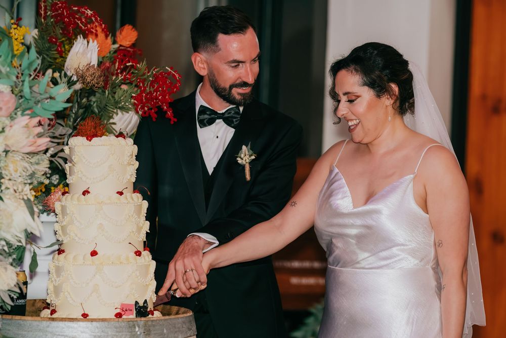 A couple cuts their elegant white wedding cake decorated with red florals and greenery during their reception.